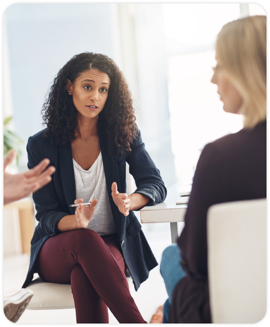 A woman with curly hair sits in an office, wearing a blazer and gesturing, engaged in a serious discussion with two others,