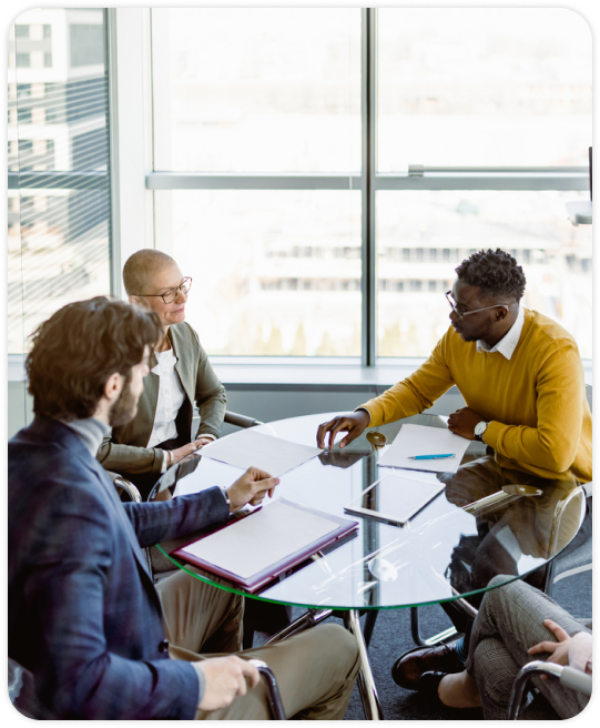 A diverse group of four professionals engaged in a meeting around a glass table in a modern office with large windows, sharing ideas and smiling.